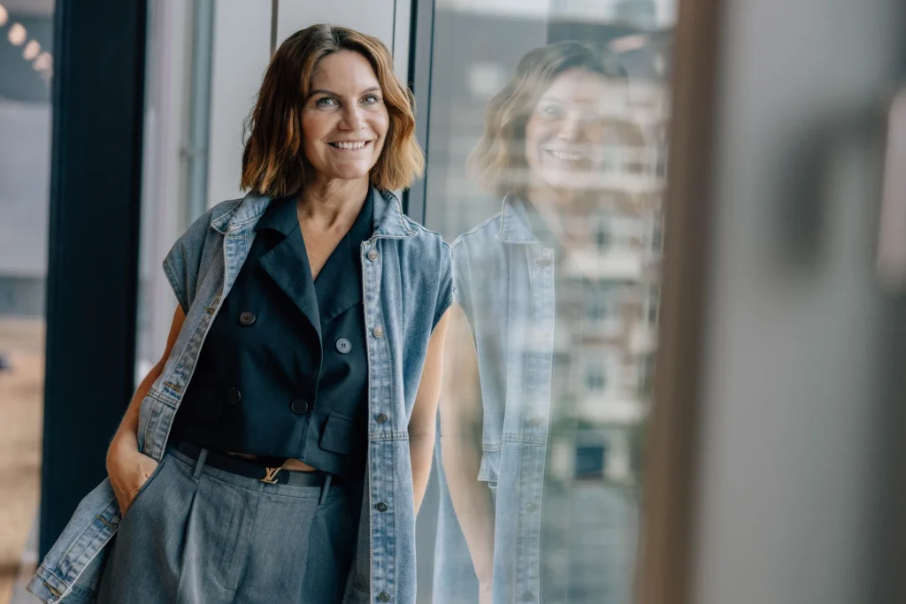 Mathilde Holmer smiling, standing by a large office window in a denim vest